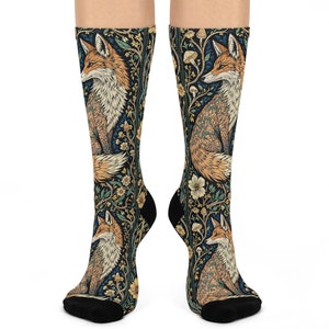 Puede incluir: Un par de calcetines con un patrón repetido de zorros y elementos florales. Los calcetines tienen un fondo azul oscuro con detalles en naranja, crema y verde. El diseño del zorro es detallado y artístico.