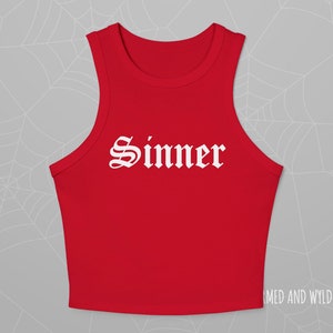 May include: A red crop top with a high neckline and the word "Sinner" printed in white gothic lettering across the front. The top is sleeveless and made of a smooth fabric.