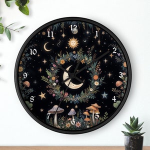 May include: A black clock with a white face and black hands. The clock face features a crescent moon, a sun, stars, and a wreath of flowers and mushrooms. The numbers on the clock face are in a white, serif font.