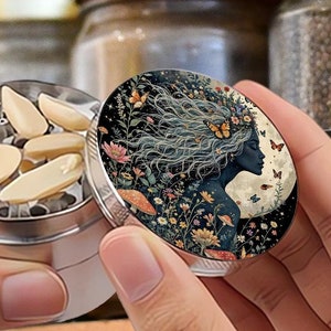 Pode incluir: Um moedor de ervas de metal prateado com uma tampa decorativa com a silhueta de uma mulher com detalhes florais e borboletas. O moedor contém amêndoas fatiadas. Frascos de especiarias são visíveis ao fundo.