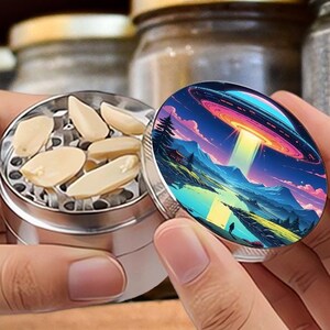 May include: A silver metal herb grinder with a colorful lid featuring a landscape with a UFO. The grinder is open, revealing the ground herbs inside. The grinder is held in front of a shelf with jars.