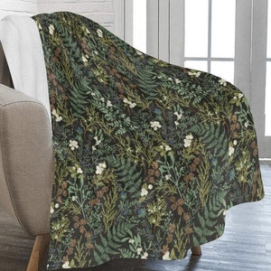 Puede incluir: Una manta de felpa con un patrón floral verde oscuro, marrón y blanco. La manta está colocada sobre un sillón de color marrón claro.