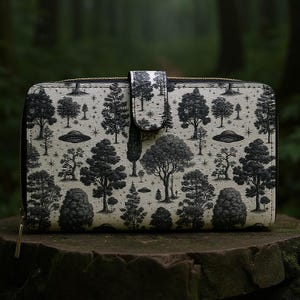 May include: A black and white wallet featuring a forest and UFO design. The wallet has a snap closure and a gold zipper. The design includes various trees and flying saucers on a light background.