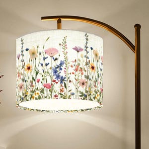 Puede incluir: Una l&aacute;mpara de pie con una pantalla floral. La pantalla tiene un fondo blanco con flores silvestres de colores en rosa, morado, amarillo y azul. La l&aacute;mpara tiene un brazo curvo de color bronce y un poste marr&oacute;n oscuro.