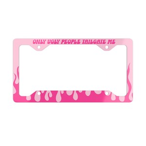 Puede incluir: Un marco de matrícula rectangular de color rosa con un patrón de llamas repetitivo en los lados y un diseño de líquido que gotea en el borde inferior. El texto "ONLY UGLY PEOPLE TAILGATE ME" está impreso en una fuente rosa en la parte superior.
