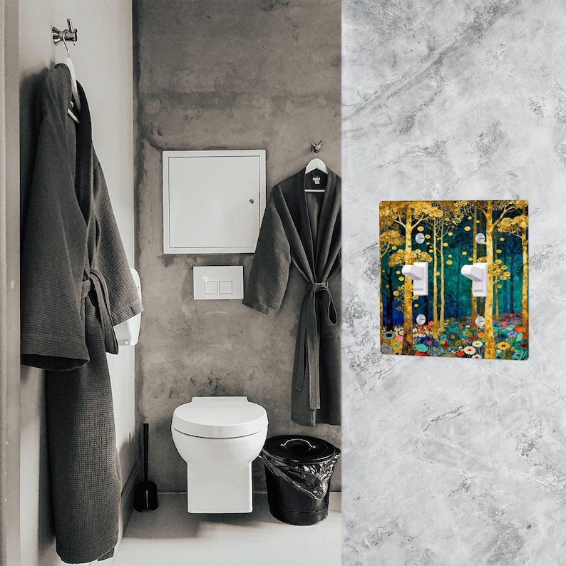 May include: A decorative light switch cover featuring a forest scene with gold, teal, and floral accents. The cover has two light switches and is mounted on a gray marble wall in a bathroom setting.