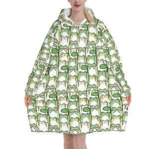 May include: A hooded blanket featuring a repeating pattern of cartoon frogs in shades of green, white, and black. The blanket has a white interior and is designed to be worn. The frog design includes lily pads.