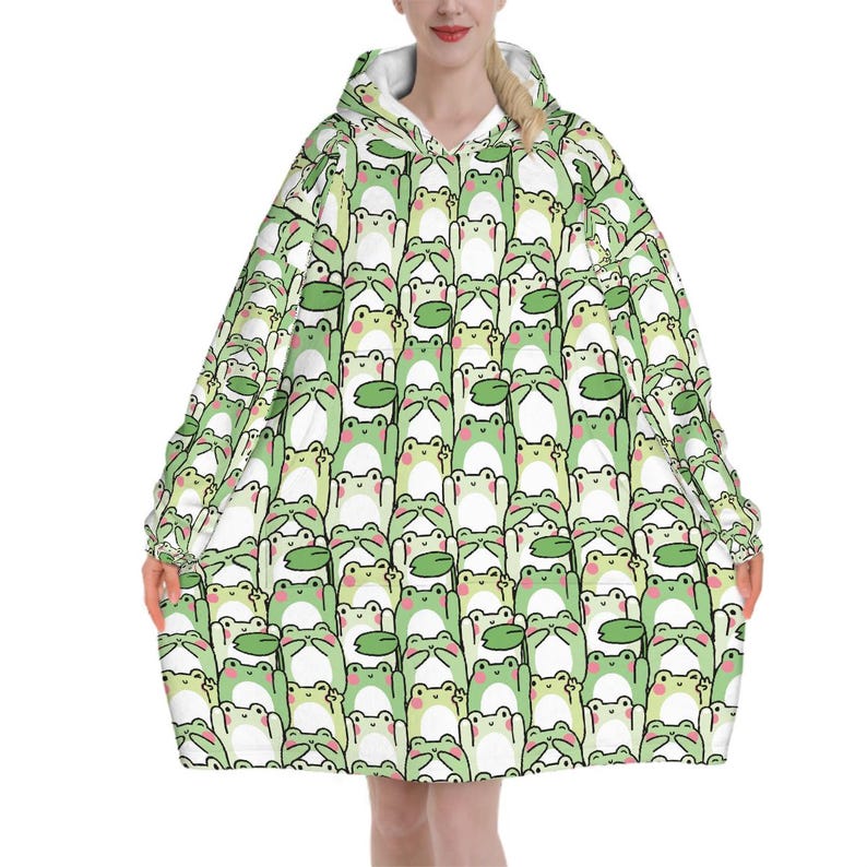 May include: A hooded blanket featuring a repeating pattern of cartoon frogs in shades of green, white, and black. The blanket has a white interior and is designed to be worn. The frog design includes lily pads.