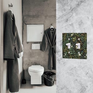 May include: A bathroom scene featuring a dark green robe hanging on a hook, a white toilet, and a black trash can. A decorative light switch cover with a floral pattern is mounted on the wall. The walls are gray.
