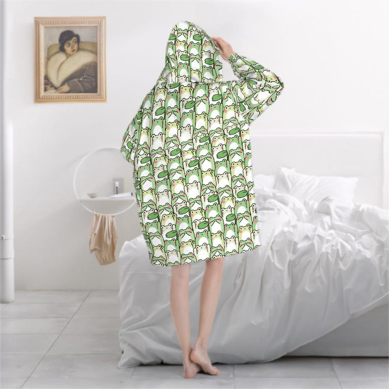 May include: A hooded blanket featuring a repeating pattern of cartoon frogs in shades of green, white, and black. The blanket is oversized and covers the person from head to mid-thigh. The background includes a white bed and a framed painting.