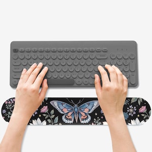 May include: A gray wireless keyboard with a matching wrist rest. The wrist rest features a black background with a large butterfly design in shades of blue, pink, and orange, surrounded by floral elements. The keyboard is being used.
