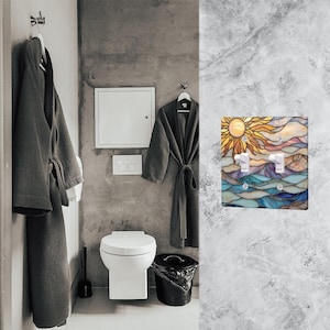 May include: A decorative light switch cover with a stained glass design depicting a sun, mountains, and water. The cover has two switches. The bathroom setting includes gray robes, a toilet, and a bin.
