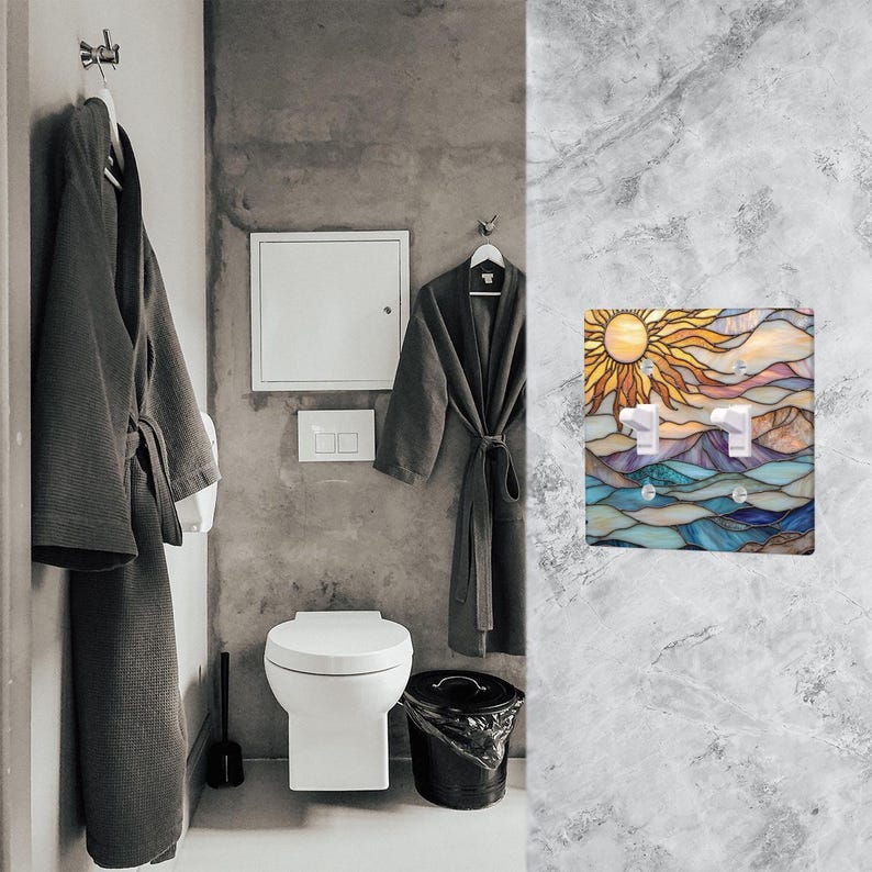 May include: A decorative light switch cover with a stained glass design depicting a sun, mountains, and water. The cover has two switches. The bathroom setting includes gray robes, a toilet, and a bin.