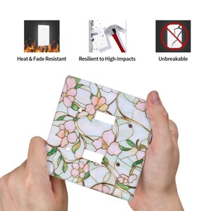 May include: A decorative light switch cover featuring a floral stained glass design with pink flowers and green leaves. The cover is held in hands and is labeled as heat & fade resistant, resilient to high impacts, and unbreakable.