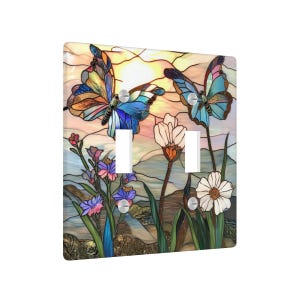 Puede incluir: Una cubierta decorativa para interruptor doble con un dise&ntilde;o de vidriera. La obra de arte representa dos mariposas con alas azules y amarillas, flores y un fondo de puesta de sol. La cubierta tiene dos aberturas para interruptores y tornillos plateados.