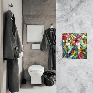 Puede incluir: Una cubierta de interruptor de luz decorativa con un dise&ntilde;o de vidriera que representa p&aacute;jaros y flores en colores vibrantes. La cubierta tiene dos interruptores y est&aacute; montada en una pared de m&aacute;rmol gris. El dise&ntilde;o incluye tonos rosas, rojos, azules y amarillos.
