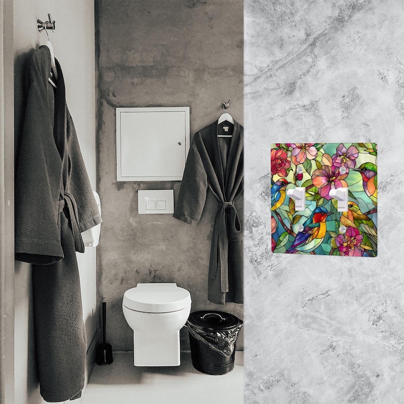 Puede incluir: Una cubierta de interruptor de luz decorativa con un dise&ntilde;o de vidriera que representa p&aacute;jaros y flores en colores vibrantes. La cubierta tiene dos interruptores y est&aacute; montada en una pared de m&aacute;rmol gris. El dise&ntilde;o incluye tonos rosas, rojos, azules y amarillos.