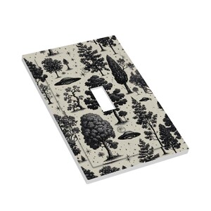 Op de afbeelding: Een lichtbeige enkele lichtschakelaar met een zwart-wit bos- en UFO-patroon. Het ontwerp toont verschillende boomsoorten, sterren en vliegende schotels. De schakelaaropening is rechthoekig.