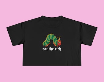 Può includere: Top corto nero con un bruco dei cartoni animati verde, giallo e rosso. Il bruco è sopra il testo "eat the rich" in un font gotico. Sfondo rosa.