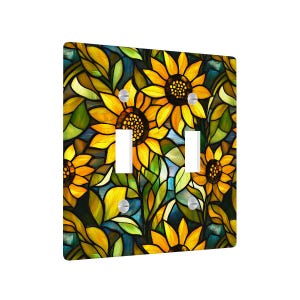 May include: A double light switch cover featuring a stained-glass design of sunflowers. The cover has a square shape with two rectangular cutouts for the switches. The sunflowers are yellow with brown centers, surrounded by green leaves and a blue background.