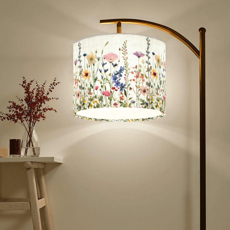 Puede incluir: Una l&aacute;mpara de pie con una pantalla floral. La pantalla tiene un fondo blanco con flores silvestres de colores en rosa, morado, amarillo y azul. La l&aacute;mpara tiene un brazo curvo de color bronce y un poste marr&oacute;n oscuro.