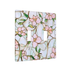 May include: A decorative light switch cover featuring a stained glass design. The cover has a white background with pink flowers, green leaves, and gold-colored outlines. The cover has two switch openings.