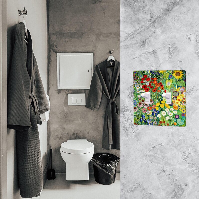 Puede incluir: Interior de ba&ntilde;o con una cubierta de interruptor de luz decorativa. La cubierta muestra un vibrante dise&ntilde;o floral con flores rojas, amarillas y azules. Dos albornoces grises cuelgan de ganchos. Un inodoro blanco y un cubo de basura negro tambi&eacute;n est&aacute;n presentes.