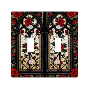 May include: A decorative double light switch cover featuring a gothic stained-glass design. The black frame is adorned with red roses and green leaves. The stained-glass panels have a cream and tan background. The light switches are silver.