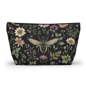 Könnte beinhalten: Schwarze Kosmetiktasche mit floralem und Bienen-Design. Die Tasche zeigt eine große Bienenillustration, umgeben von verschiedenen Blumen und Blättern in Creme-, Rosa- und Grüntönen. Der Reißverschluss ist goldfarben.