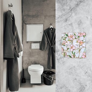 May include: A bathroom scene featuring a white toilet, a black trash can, and two gray bathrobes hanging on hooks. A decorative light switch cover with a floral design in pink, green, and white adds a touch of color.
