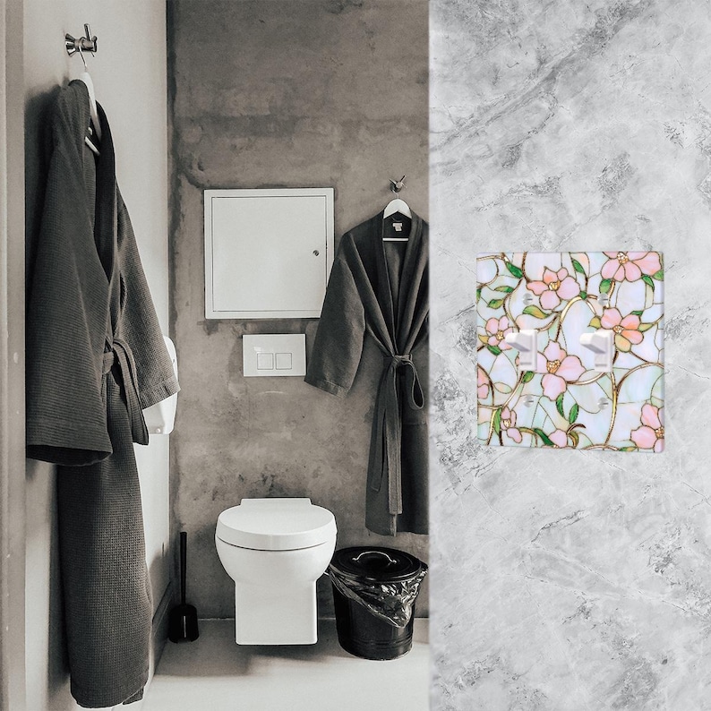 May include: A bathroom scene featuring a white toilet, a black trash can, and two gray bathrobes hanging on hooks. A decorative light switch cover with a floral design in pink, green, and white adds a touch of color.