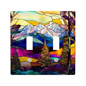 Puede incluir: Una cubierta decorativa para interruptor de luz doble con un diseño de vidriera de un paisaje montañoso. La obra de arte incluye un cielo colorido, montañas nevadas y árboles en tonos de azul, morado, amarillo y naranja. La cubierta es cuadrada con dos aberturas para interruptores.