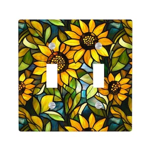 Puede incluir: Cubierta de interruptor de luz doble con un diseño de girasoles de vidrieras. La cubierta cuadrada tiene dos recortes para los interruptores. Los girasoles son amarillos con centros marrones, sobre un fondo de hojas verdes y azul.