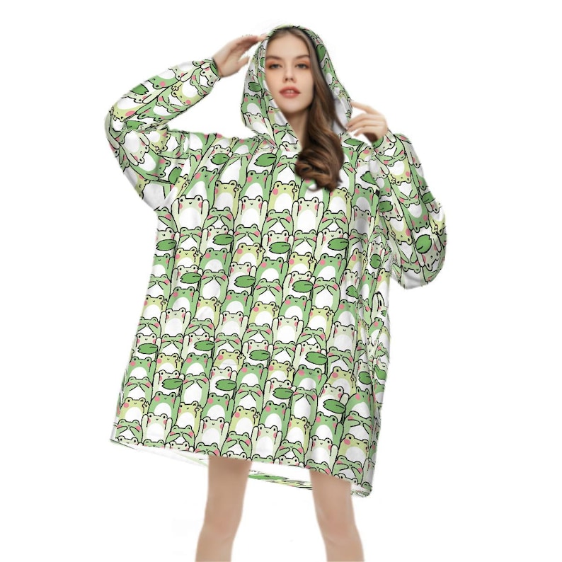 May include: A hooded blanket featuring a repeating pattern of cartoon frogs in shades of green, white, and pink. The blanket has long sleeves and a hood, offering a cozy and whimsical design. The frog pattern is consistent across the entire blanket.