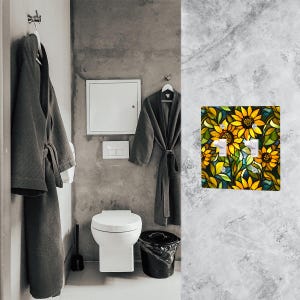 May include: A bathroom scene featuring a decorative light switch cover with a sunflower design. The cover has two switches and is surrounded by yellow, green, and blue floral elements. The bathroom also includes a toilet, a trash can, and two gray bathrobes.