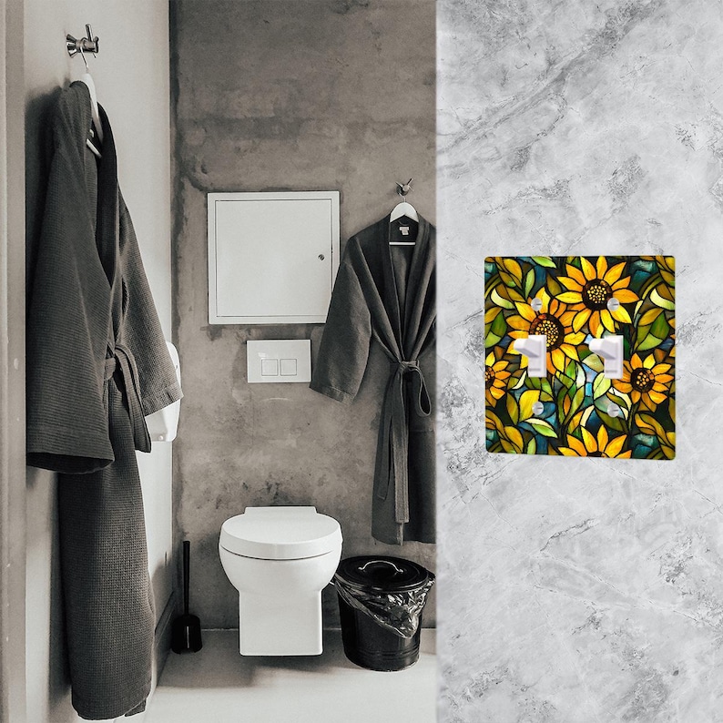 May include: A bathroom scene featuring a decorative light switch cover with a sunflower design. The cover has two switches and is surrounded by yellow, green, and blue floral elements. The bathroom also includes a toilet, a trash can, and two gray bathrobes.