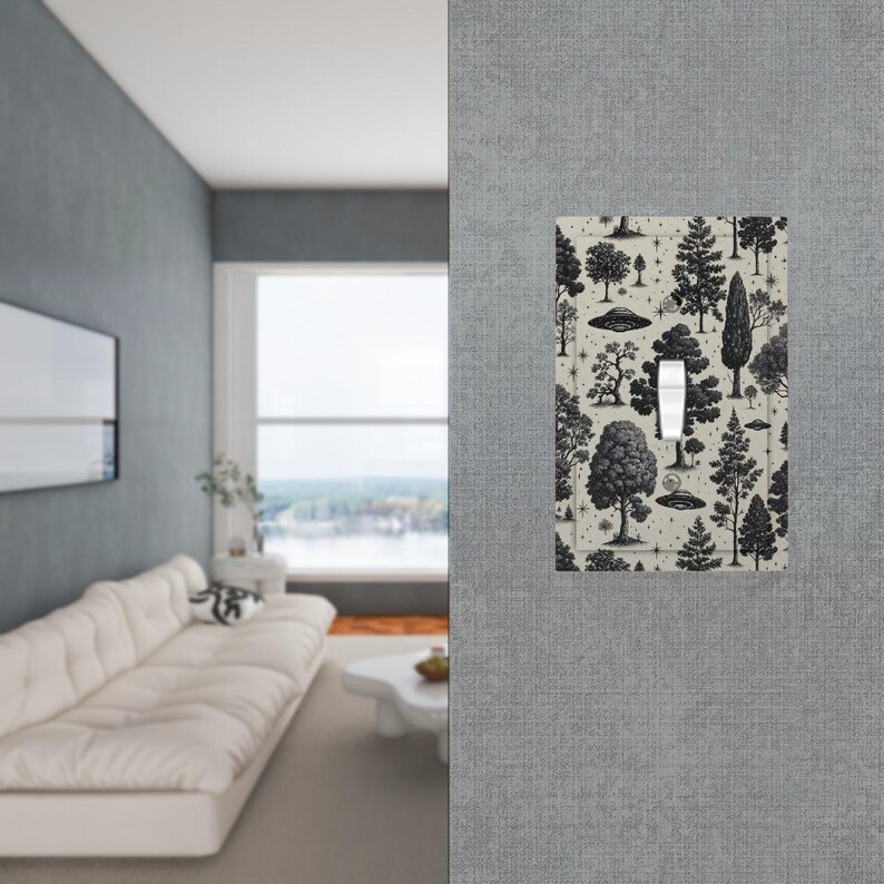 Op de afbeelding: Een lichtschakelaar met een zwart-wit bospatroon met bomen en UFO's. De schakelaar staat in de aan-stand. De afdekking is op een grijze muur gemonteerd.