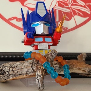 Op de afbeelding: Een rood, blauw en zilveren speelgoedrobot, Optimus Prime, zit op een tak met een blauwe en oranje kralenarmband. De robot houdt een geel en oranje zwaard vast.