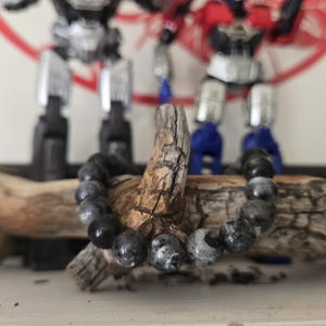 Op de afbeelding: Een armband van zwarte en grijze kralen met een natuurlijke tak op de achtergrond.