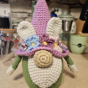 May include: A crocheted gnome with a purple hat, white beard, and green body. The gnome is wearing a flower crown with pink, purple, blue, and yellow flowers.