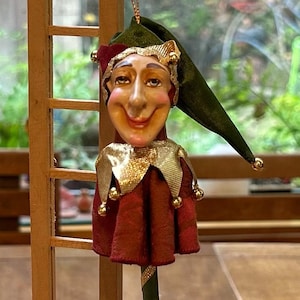 May include: A whimsical jester ornament with a smiling face, wearing a green hat and a red and gold outfit. The jester's staff is green with gold accents. The ornament is hanging near a small wooden ladder.