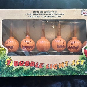 Könnte beinhalten: Ein 7er-Set Blasenlichter in einem rechteckigen Karton mit orangefarbenen kürbisförmigen Sockeln und Jack-o'-Lantern-Gesichtern. Die Verpackung enthält den Text 7 BUBBLE LIGHT SET, See Them Bubble! und UL LISTED sowie Abbildungen von grünen Tannenzweigen.