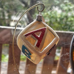 Peut inclure: Une décoration de Noël transparente en forme de cube, avec un "A" rouge pailleté sur un côté et un "B" bleu pailleté sur un autre. L'ornement a une bordure dorée et est suspendu par un fil doré. L'arrière-plan est une vue floue d'une fenêtre.
