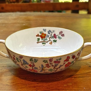 以下が含まれることがあります： 赤、オレンジ、青、ピンクの花柄が施された、2つの取っ手が付いた白いセラミック製のスープボウル。縁は金でトリミングされています。ボウルは木製の表面に置かれています。