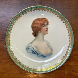 Puede incluir: Plato decorativo de porcelana con un retrato de una mujer pelirroja con un vestido azul. El plato tiene un fondo blanco con un borde decorativo verde y dorado. La mujer mira hacia la derecha.