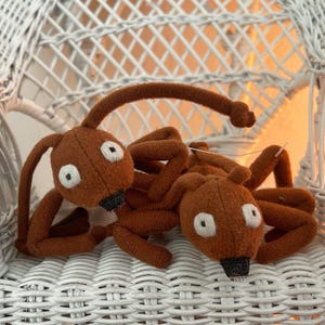 May include: Two brown felt insect toys with white eyes and black noses are resting on a white wicker chair. The toys have long, thin legs and antennae. The toys are handmade and have a whimsical design.