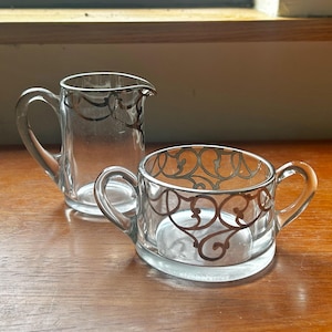 Może przedstawiać: Przezroczysty szklany zestaw do śmietanki i cukru. Oba elementy mają srebrny wzór filigranowy wokół krawędzi i po bokach. Śmietanka ma uchwyt i wylewkę. Cukiernica ma dwa uchwyty.