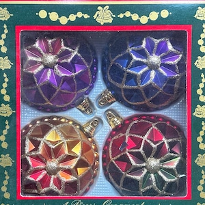 Könnte beinhalten: Ein Set aus vier Weihnachtskugeln in einer Schachtel. Die Kugeln sind juwelenfarben mit goldenen Akzenten und Sternenmustern. Die Schachtel ist grün mit rotem Rand und dekorativen Elementen.