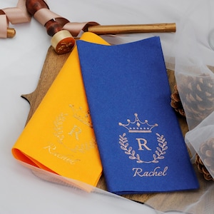 May include: Two paper napkins with a gold crown and the letter R in a wreath design. The napkins are blue and yellow and have the name Rachel printed on them.