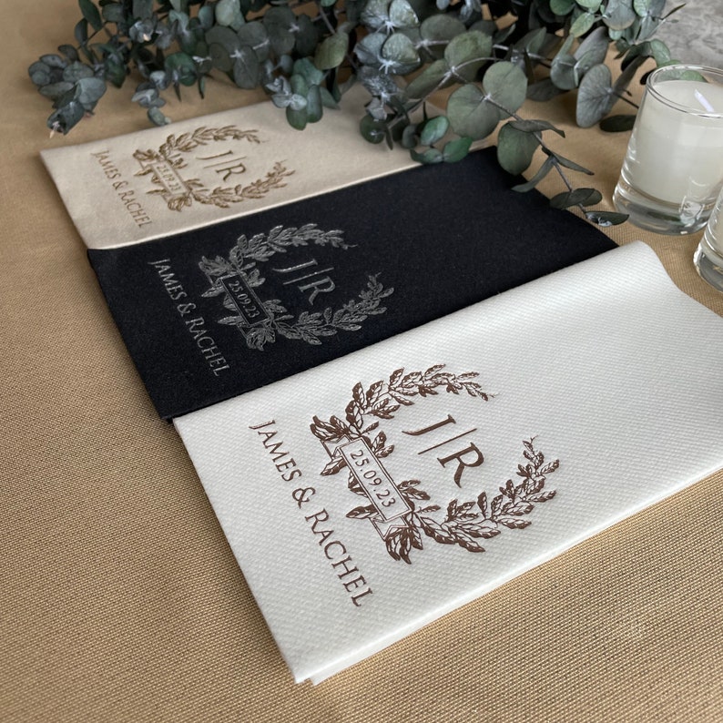 May include: Three white linen napkins with a brown monogram and the text "J/R 25.09.23" and "JAMES & RACHEL". One napkin is black with a silver monogram.